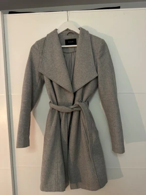 Ljusgrå kappa från Vila med skärp - Stilren figursydd ljusgrå kappa från Vila med både knappar och knytskärp i midjan. Tllverkad i ett mjukt tyg som ger en klassisk look. Perfekt för dig som gillar minimalistisk och trendig stil. Slutar strax över knäna på mig som är 175!❤️
