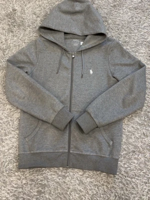 Grå hoodie från Polo Ralph Lauren - Snygg grå hoodie från Polo Ralph Lauren med dragkedja framtill, huva med dragsko och två fickor. Klassisk logga broderad på bröstet i vitt. Tillverkad i mjuk bomullsblandning som känns skön mot huden. Perfekt för en avslappnad och stilren look.
