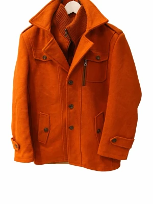 Orange ullrock med dragkedja och knappar - Snygg orange rock i ull med bred krage, knappar framtill och dragkedja vid bröstet. Jackan har flera fickor, ribbad hög krage på insidan och detaljer vid ärmslut. Perfekt i höst och vinter. Det är även inte så skrikig orange och kragen sitter med dragkedjan på insidan så den gör även att ta bort. Det ör en XL men skulle säga att den sitter som en L/M 