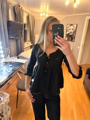 Svart blus med rosett och spetsdetaljer - Pris kan diskuteras. Svart blus med lång ärm, v-ringning och knytning framtill. Dekorerad med en liten röd rosett under bysten och diskreta spetsdetaljer på bröstet. Blusen har en lätt utsvängd passform och är tillverkad i ett tunt, mjukt tyg.