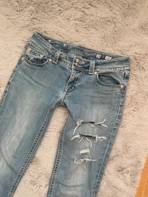 Miss Me ljusblå low waist bootcut jeans med slitningar - Säljer ett par ljusblå jeans från Miss Me med bootcut passform och snygga slitningar på ena benet. Jeansen har dekorativa bakfickor med broderade detaljer och pärlknappar. Klassisk femficksmodell i jeansmaterial med låg midja. Har sytt längst in i byxan men annars jättebra skick