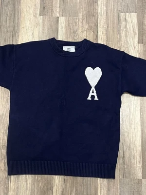 Mörkblå stickad tröja från Ami - Mörkblå stickad tröja från Ami med rund halsringning och ribbade muddar. På bröstet finns ett vitt hjärta ovanför ett stort A, vilket är varumärkets ikoniska logga. Perfekt för dig som gillar stilrena och trendiga plagg.