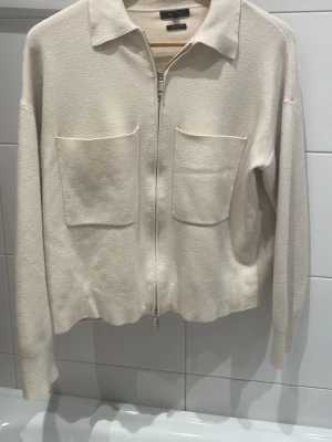 Beige cardiganjacka Massimo Dutti - Stilren beige cardiganjacka från Massimo Dutti med dragkedja framtill, två stora bröstfickor och klassisk krage. Jackan har lång ärm och är tillverkad i ett mjukt, strukturerat material som ger en clean look.