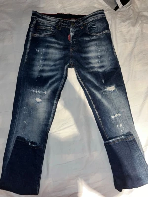 Dsquared2 slitna blå jeans - Säljer ett par blå jeans från Dsquared2 med slitningar, hål och patchade detaljer. Jeansen, låg midja och klassisk knappgylf. Snyggt tvättade med en cool, använd look och Dsquared2-märke bak i midjan.