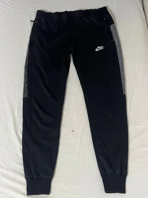 Svarta Nike mjukisbyxor med vita detaljer - Svarta mjukisbyxor från Nike med vita prickade detaljer längs sidorna och klassisk Nike-logga på låret. Byxorna har resår i midjan och muddar vid bensluten. Perfekta för chill eller träning. Har ett jättelitet hål vid ena knäna. Saknar band. Storleken på lappen är XXL men i verkligheten är den M/L.