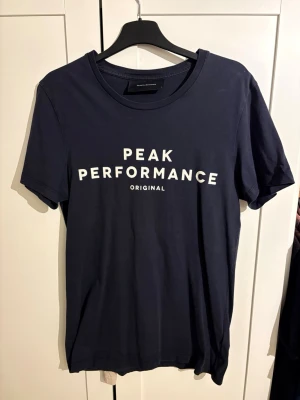 Mörkblå Peak Performance t-shirt - Snygg mörkblå t-shirt från Peak Performance med vit text på bröstet. Klassisk rund halsringning och korta ärmar. Tillverkad i mjuk bomull som känns skön mot huden. Perfekt för en avslappnad och stilren look.
