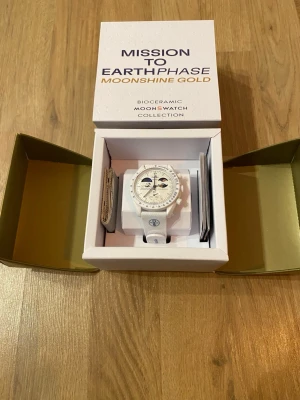 Swatch x Omega MoonSwatch Earthphase - Snygg vit Swatch x Omega MoonSwatch från Bioceramic MoonSwatch Collection, modellen Mission to Earthphase Moonshine Gold. Klockan har vit boett och armband, samt detaljer i guld och blått på urtavlan. Kommer i originalförpackning.