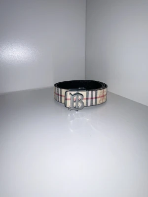 Randigt Burberry bälte med B-spänne - Snyggt bälte man kan byta sida på den så man väljer melan en brun aktig färg eller en svart aktig färg. Deta bältet är andvänt ca 1 gång och det var till ett bröllop