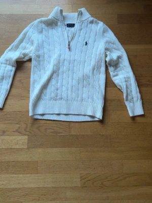 🥥Vit kabelstickad tröja Polo Ralph Lauren - Kabelstickad vit tröja från Polo Ralph Lauren med halv dragkedja och klassisk broderad logga på bröstet. Tröjan har ribbade muddar och krage, samt är tillverkad i mjukt material som ger en lyxig känsla.