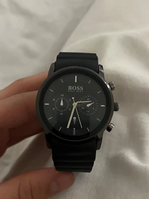 Svart klocka från Hugo Boss - Stilren svart klocka från Hugo Boss med svart silikonarmband och rund urtavla. Klockan har silverfärgade detaljer, två mindre urtavlor och knappar på sidan. Baksidan är i rostfritt stål. Perfekt accessoar för en clean look.