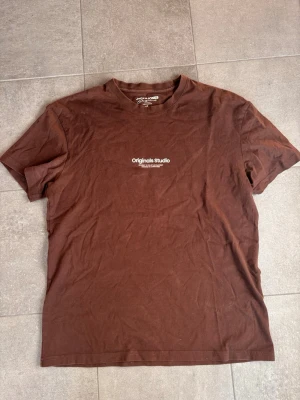 Brun t-shirt från Jack & Jones - Snygg brun t-shirt från Jack & Jones med trycket 'Originals Studio' på bröstet. Klassisk rund hals och korta ärmar, perfekt för en avslappnad stil. Tillverkad i mjuk bomull som känns skön mot huden.