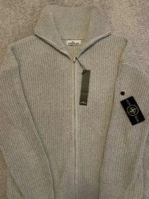 Grå stickad ziptröja från Stone Island - Snygg grå stickad tröja från Stone Island med dragkedja framtill och hög krage. Tröjan har ribbstickad struktur och klassisk Stone Island-logga på ärmen. Perfekt för lager-på-lager och stilren look. Har i  grå färg och svart färg