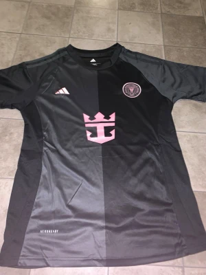 Svart Inter Miami CF matchtröja Adidas - Snygg svart matchtröja från Inter Miami CF med rosa detaljer och klubbmärke på bröstet. Tröjan har Adidas-logga, AEROREADY-material och en modern passform. Perfekt för fotbollsträning eller att bära som supporter.