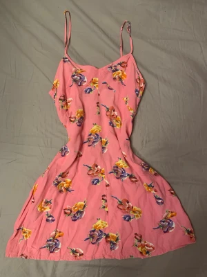 Rosa blommig sommarklänning - Söt rosa klänning med smala axelband och färgglatt blommönster i lila, gult och orange. Klänningen är kort och har en lätt utsvängd passform, perfekt för varma dagar. Dragkedja i ryggen för enkel påklädning.