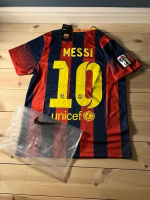 FC Barcelona Messi matchtröja Nike - Säljer en FC Barcelona matchtröja från Nike med Messi 10-tryck på ryggen. Tröjan har röda och blå vertikala ränder, gula detaljer och klubbmärke fram. Materialet är lätt och andas, perfekt för fotboll eller träning. La Liga-märke på ärmen.