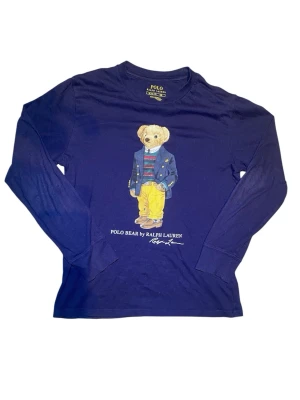 Blå långärmad Polo Bear t-shirt - Snygg mörkblå tunn långärmad t-shirt från Ralph Lauren med ikoniska Polo Bear-trycket framtill. Klassisk rund hals och mjukt bomullsmaterial som känns skönt mot huden. Perfekt för dig som gillar streetstyle och vill ha något som sticker ut.
