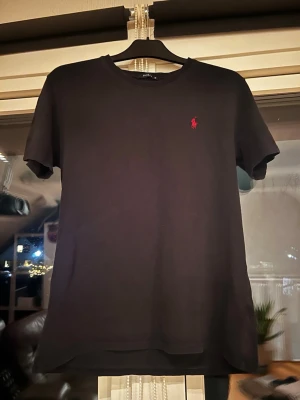 Svart t-shirt från Polo Ralph Lauren - Klassisk svart t-shirt från Polo Ralph Lauren med röd broderad logga på bröstet. T-shirten har rund halsringning och korta ärmar. Tillverkad i mjuk bomull för en skön känsla och enkel stil.