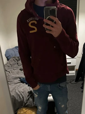 Vinröd hoodie från Hollister - Snygg vinröd hoodie från Hollister med tryckt logga och bokstaven S i gult på bröstet. Klassisk huva och känguruficka framtill. Perfekt för en avslappnad och trendig stil.