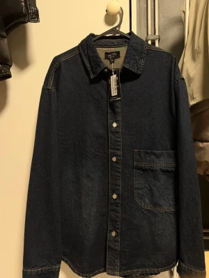 Mörkblå jeansjacka från MAN - Stilren mörkblå jeansjacka från MAN med klassisk krage, knappstängning framtill och en stor bröstficka på vänster sida. Jackan har raka ärmar och kontrastsömmar som ger en snygg detalj. Perfekt för dig som gillar enkel och tidlös denimstil.