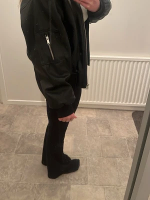 Svart bomberjacka med fickdetaljer - Säljer en svart bomberjacka med dragkedja framtill och ficka med dragkedja på ärmen. Jackan har en klassisk oversized passform och ribbade muddar vid ärmslut och midja. Perfekt för dig som gillar en streetwear-look.