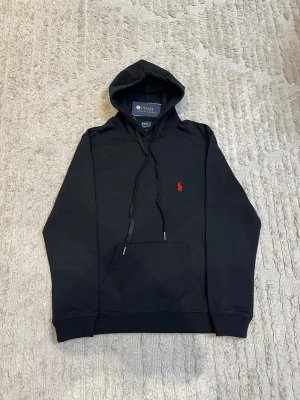 Svart hoodie från Polo Ralph Lauren - Svart hoodie från Polo Ralph Lauren med klassisk huva och dragsnören. Har en röd broderad logga på bröstet och en stor magficka framtill. Tillverkad i mjukt bomullsmaterial, perfekt för en avslappnad och stilren look.