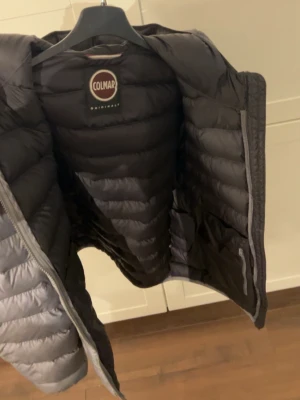 Grå dunjacka med huva från Colmar - Snygg grå dunjacka från Colmar med huva och quiltad design. Jackan har dragkedja framtill och är lätt vadderad för extra värme. Perfekt för kalla vinterdagar och har en modern, sportig look. Tillverkad i ett slitstarkt material med klassisk Colmar-logga på insidan.