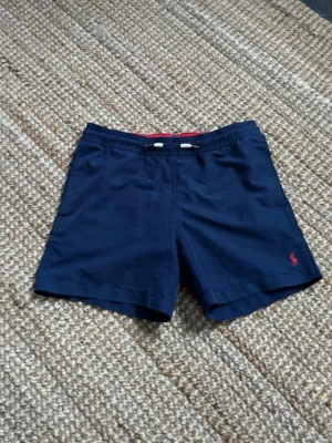 Marinblå badbyxor Polo Ralph Lauren - Marinblå badbyxor från Polo Ralph Lauren med röd broderad logga på benet. Klassisk modell med resårmidja och vit dragsko samt röd insida i midjan. Tillverkade i snabbtorkande material, perfekta för strand och pool.