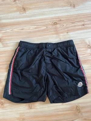 Svarta MoncIer badbyxor med ränder - Snygga svarta badbyxor från MoncIer med röda, vita och blå ränder längs sidorna. Klassisk MoncIer-logga på vänster ben. Elastisk midja med knappstängning och lätt, snabbtorkande material. Perfekta för stranden eller poolen.