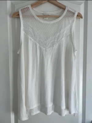 Vit ärmlös topp med spetsdetaljer - Säljer en vit ärmlös topp från H&M med transparent mesh upptill och fina spetsdetaljer över bröstet. Toppen har en mjuk och luftig känsla, perfekt för varma dagar. Passar till både jeans och kjol för en fräsch look.