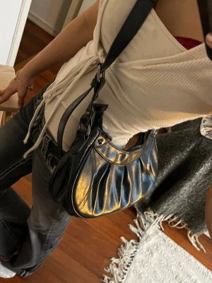 Svart axelväska i skinnimitation - Trendig svart axelväska med justerbar bred rem och snygga metalldetaljer. Väskan har en rundad form och dragkedja upptill. Materialet är glansig skinnimitation som ger en cool look. Perfekt för dig som gillar en stilren men edgy accessoar.