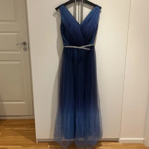 Blå glittrig långklänning med v-ringning - Säljer en magisk blå långklänning med v-ringad omlott-topp och glittrande detaljer. Klänningen har ett tunt lager tyll med diskret glitter och ett snyggt bälte i midjan med strass. Perfekt för bal eller fest! Snyggt fall och ärmlös design.