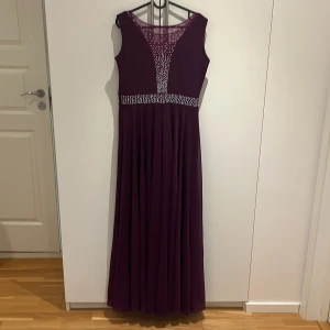 Lång vinröd klänning med paljettdetaljer - Lång vinröd klänning med ärmlös design och v-ringad mesh upptill. Klänningen har glittrande paljettdetaljer längs midjan och bröstet, vilket ger en elegant look. Flödande kjol i lätt material, perfekt för speciella tillfällen.