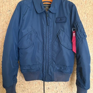 Blå flygarjacka från Alpha Industries - Vadderad, blå nylonjacka från Alpha Industries i herrstorlek Medium. Modellen heter ”Flyer’s CWU VF TT”. Mycket sparsamt använd och kommer från ett djur- och rökfritt hem.