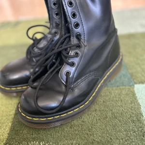 Svarta Dr. Martens boots i skinn - Klassiska svarta Dr. Martens boots i skinn med snörning och ikonisk gul söm runt sulan. Grov, genomskinlig sula med bra grepp och högre skaft. Perfekta för dig som vill ha en tidlös och edgy stil.