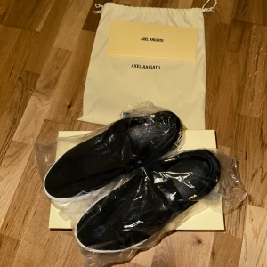  Axel Arigato laceless - Svarta slip-on sneakers från Axel Arigato med ovandel i mocka och elastiskt band över vristen. Skorna har vit platt sula och minimalistisk design, perfekt för en clean och stilren look. Levereras med dustbag och originalkartong. Använda 1 gång för ett bröllop