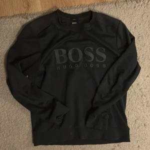 Svart slim fit tröja från Hugo Boss - Svart långärmad tröja från Hugo Boss med stort BOSS-tryck på bröstet. Slim fit-modell med ribbade muddar vid ärmslut och nederkant. Tillverkad i mjuk bomull för en skön känsla och stilren look.