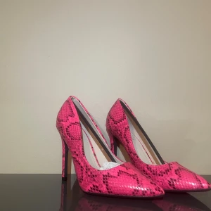Rosa pumps med ormskinnsmönster - Säljer ett par riktigt snygga pumps i starkt rosa med ormskinnsmönster. Skorna har spetsig tå och hög klack, vilket ger en edgy och trendig look. Materialet är glansigt syntet som ger extra attityd. Perfekta för dig som vill sticka ut och älskar färg!