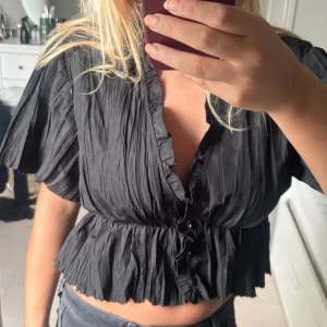 Svart croppad blus med volang - Säljer en svart croppad blus med korta ärmar och v-ringning. Blusen har volangdetaljer längs halsringningen och nederkanten samt knappar framtill. Materialet är lätt och har en snygg skrynklig struktur som ger en cool vibe.