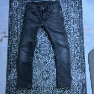 Svarta jeans från Nudie Jeans, W31 L30 - Säljer ett par svarta jeans från Nudie Jeans med klassisk femficksdesign och broderade bakfickor. Jeansen har en smal passform och är tillverkade i mjukt denimtyg. Perfekta för dig som gillar stilrena och bekväma jeans. Liten lagning profetionellt gjord av nudiejeans i grenen sista bilden. Hör av dig vid frågor eller prisförslag!!