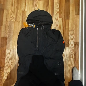 Svart vinterjacka Parajumpers herr L - Grym svart vinterjacka från Parajumpers med huva, coola detaljer som orange rem vid kragen och patch på ärmen. Jackan har flera fickor, dragkedja och knappar framtill. Perfekt för kalla dagar och riktigt snygg streetstil.