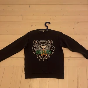 Svart Kenzo sweatshirt med tigertryck XS - Svart sweatshirt från Kenzo i storlek XS med ikoniskt broderat tigerhuvud och färgglada detaljer framtill. Tröjan har rund halsringning och långa ärmar. Perfekt statement-plagg för dig som vill sticka ut.