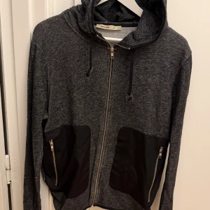 Mörkgrå hoodie från Whyred med dragkedja - Snygg mörkgrå hoodie från Whyred med hel dragkedja framtill och två svarta fickor med dragkedjor. Tröjan har huva med snörning och är tillverkad i en mix av bomull, polyester och elastan. Perfekt för en chill och stilren look. Storlek M.