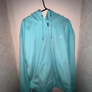 Polo Ralph Lauren Zip Hoodie - Säljer en turkos hoodie från Ralph Lauren med dragkedja, huva och snörning. Tröjan har två stora fickor framtill och den klassiska vita loggan broderad på bröstet. Perfekt för dig som gillar en chill och sportig stil.