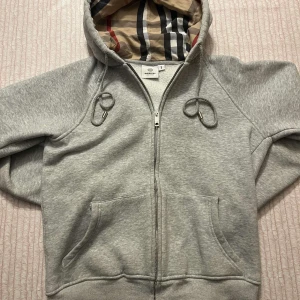 Grå hoodie från Burberry med dragkedja - Säljer en grå hoodie från Burberry med klassiskt rutigt foder i huvan. Tröjan har hel dragkedja, snörning i huvan och två stora fickor framtill. Perfekt för dig som vill ha en stilren och lyxig vardagslook.