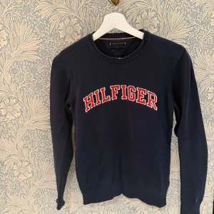 Mörkblå stickad tröja Tommy Hilfiger - Mörkblå stickad tröja från Tommy Hilfiger med stort broderat HILFIGER-tryck i rött och vitt på bröstet. Klassisk rund halsringning och ribbade muddar vid ärmslut och nederkant. Perfekt för dig som gillar preppy och stilren look.