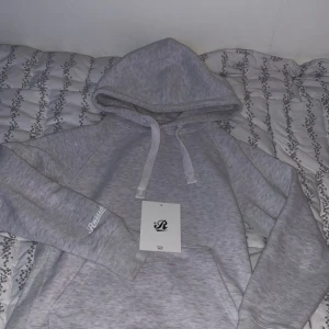  Russemerch hoodie - Ljusgrå hoodie från Russedress i storlek S. Tröjan har huva med snörning, magficka och broderad logga på ärmen. Mjuk insida och klassisk passform, perfekt för chill dagar. Enkel och stilren design som funkar till det mesta. Inga defekter eller fläckar. Köpt för 3 veckor sedan 