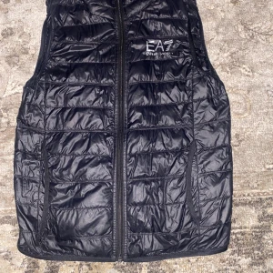 Svart dunväst från EA7 Emporio Armani - Svart quiltad dunväst från EA7 Emporio Armani med dragkedja framtill och två sidofickor. Västen har en stilren design med EA7-logga på bröstet och lätt glansigt material. Perfekt för lager-på-lager och snygg till streetwear.