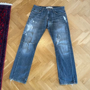 Levi's 514 straight jeans blå denim - Levi's 514 straight jeans i klassisk blå denim med slitna detaljer, färgstänk och fransade benslut. Byxorna har fem fickor, orange sömmar och en rak passform. Perfekta för dig som gillar en avslappnad och vintage-inspirerad look.