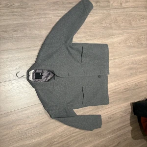 Grå kostymjacka  - Stilren grå jacka från lager 157 i ullblandning med klassisk krage och raka linjer. Jackan har två stora fickor framtill, knäppning med knappar och quiltat foder för extra komfort. Perfekt för dig som gillar minimalistisk och clean look.