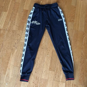 Blå träningsbyxor Better Bodies S - Mörkblå träningsbyxor från Better Bodies med vita sidor och svarta loggor längs benen. Byxorna har snörning i midjan, muddar vid bensluten och röda detaljer. Perfekta för gymmet eller chill. Materialet är mjukt och syntetiskt.